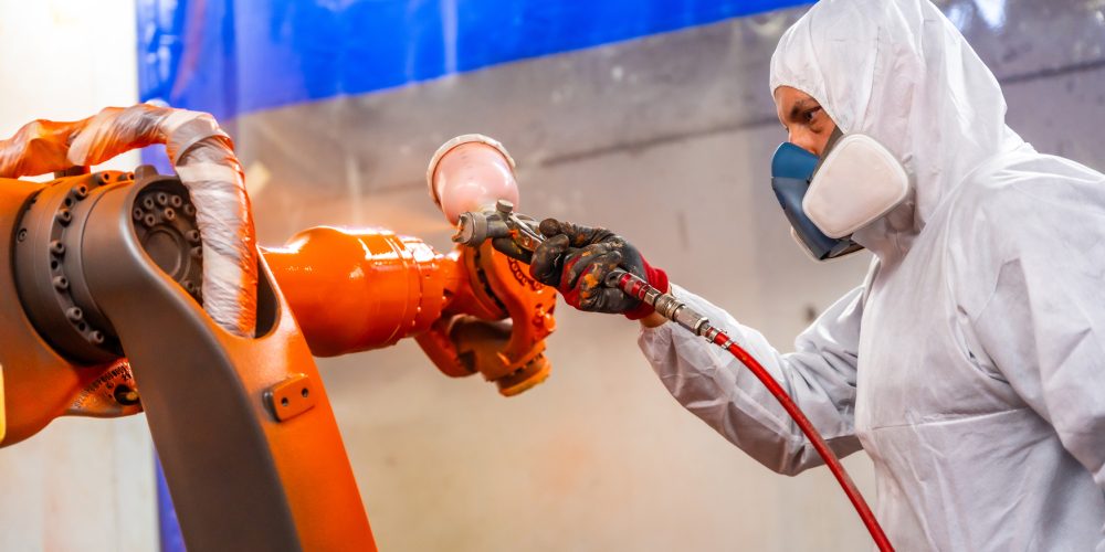 Painter with protective gear using a spray gun to paint an industrial robot arm in a factory